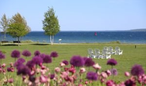 Flowers bloom in the foreground of Mission Point Resort's great lawn with white Adirondack chairs and blue water in the distance