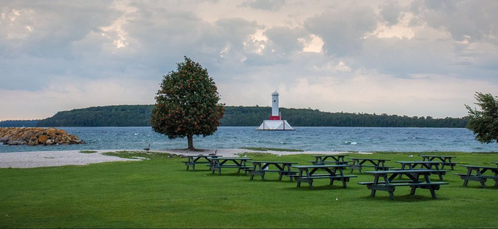 Picnic tables fill the lawn at Mackinac Island’s Windermere Point on a cloudy day with a tree and lighthouse in focus