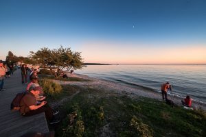 Several people sit on the Mackinac Island boardwalk while another sets up a tripod on the beach before sunset.